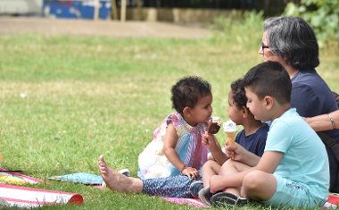 Three small children and an adult eat ice creams while sat on a picnic blanket in Barra Hall Park on a sunny day.