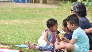 Three small children and an adult eat ice creams while sat on a picnic blanket in Barra Hall Park on a sunny day.